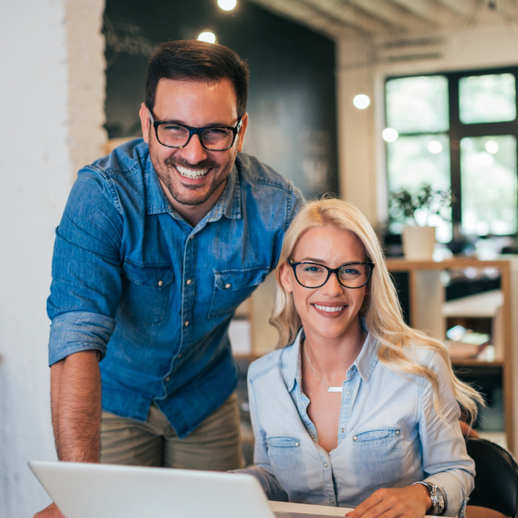 Two office workers smile at the camera. They are sitting in front of a laptop.